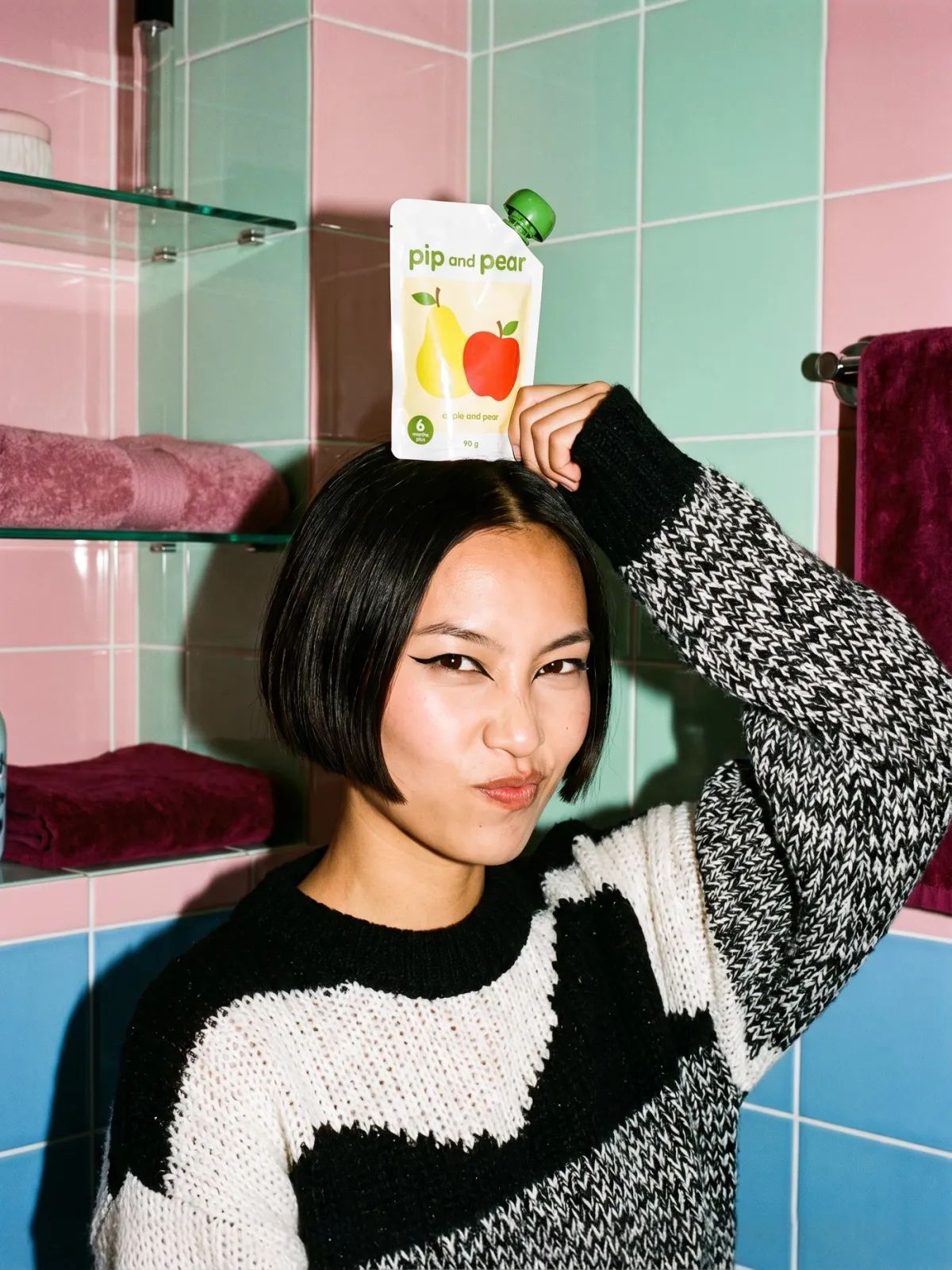 A woman balances a pip and pear pouch on her head in a pink and mint tiled bathroom