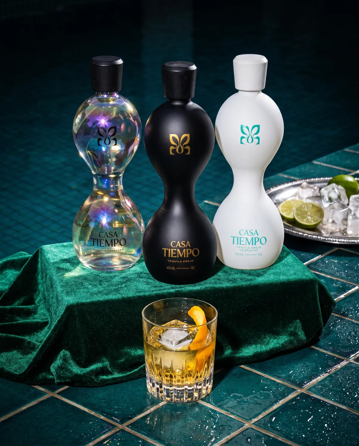 Three Casa Tiempo bottles on emerald velvet at the pool edge, with a poured rocks glass in front.